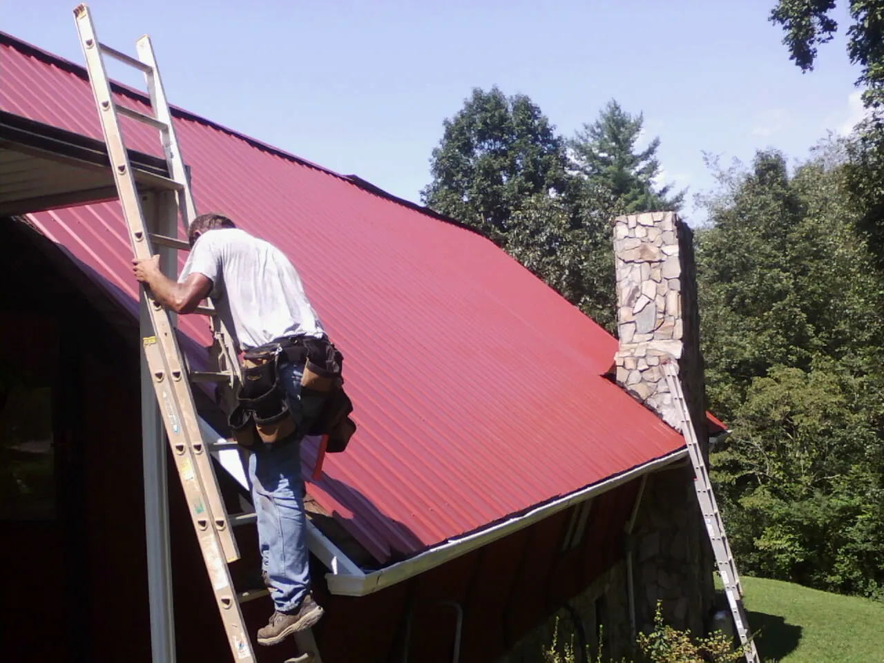 Roofer on a ladder beside a red metal roof and stone chimney — Soffit & Fascia Repair by Roofing Brothers in Laguna Hills